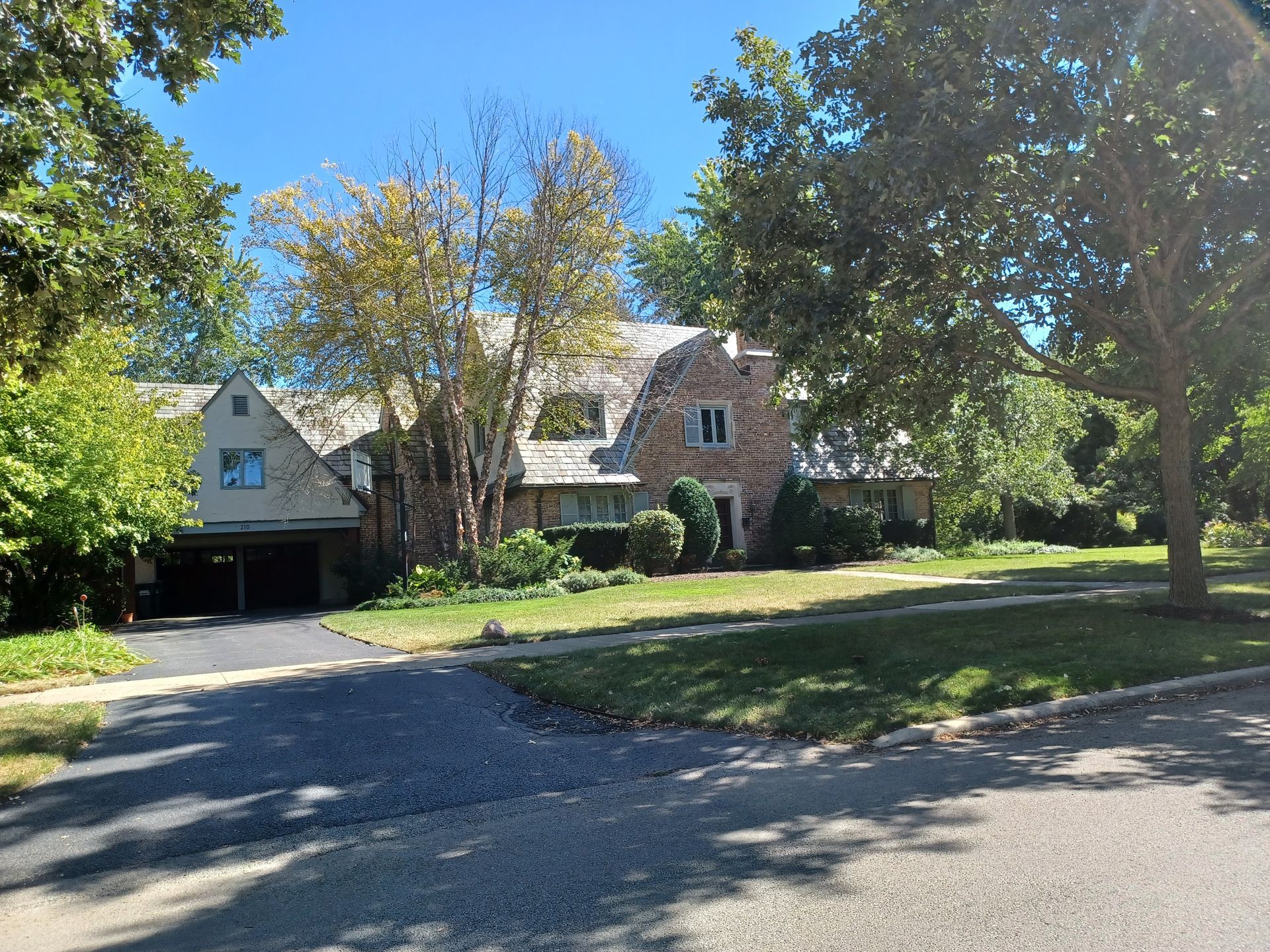 Stone house with a carport, set on a green lawn, surrounded by trees under a blue sky.