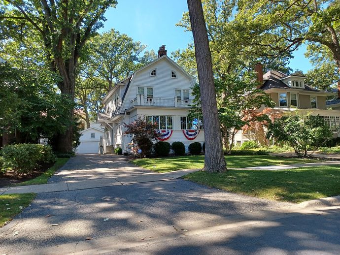White two-story house with American flag bunting; driveway on left, trees surround, sunny day.