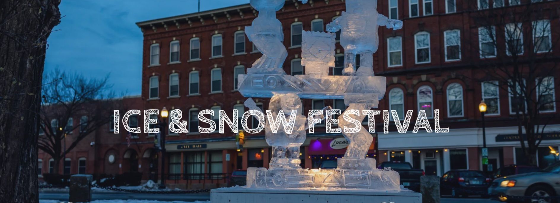 Ice and Snow Festival sign lit on a snowy street in front of brick buildings at dusk