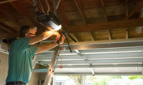 Person on a ladder repairs a garage door opener under a wooden ceiling.