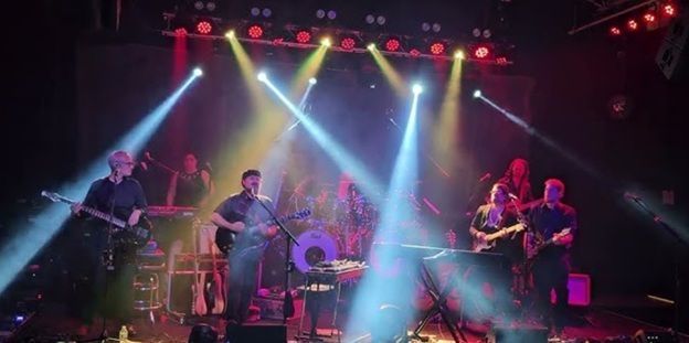 Live band performing on stage under blue and pink spotlights with drums and guitars