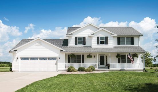 White two-story suburban house with a front porch, attached garage, and green lawn under a blue sky