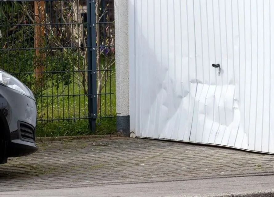 White garage door with a large dent beside a parked car and fence, on a paved driveway.