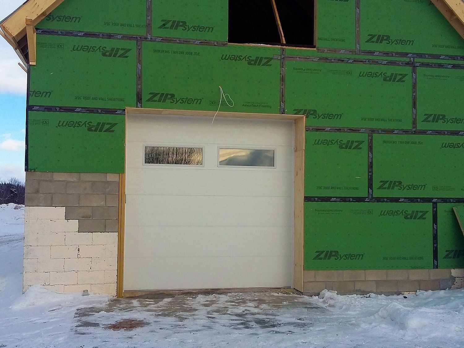 Partially finished garage wall with green insulation panels and a white overhead door in snow.