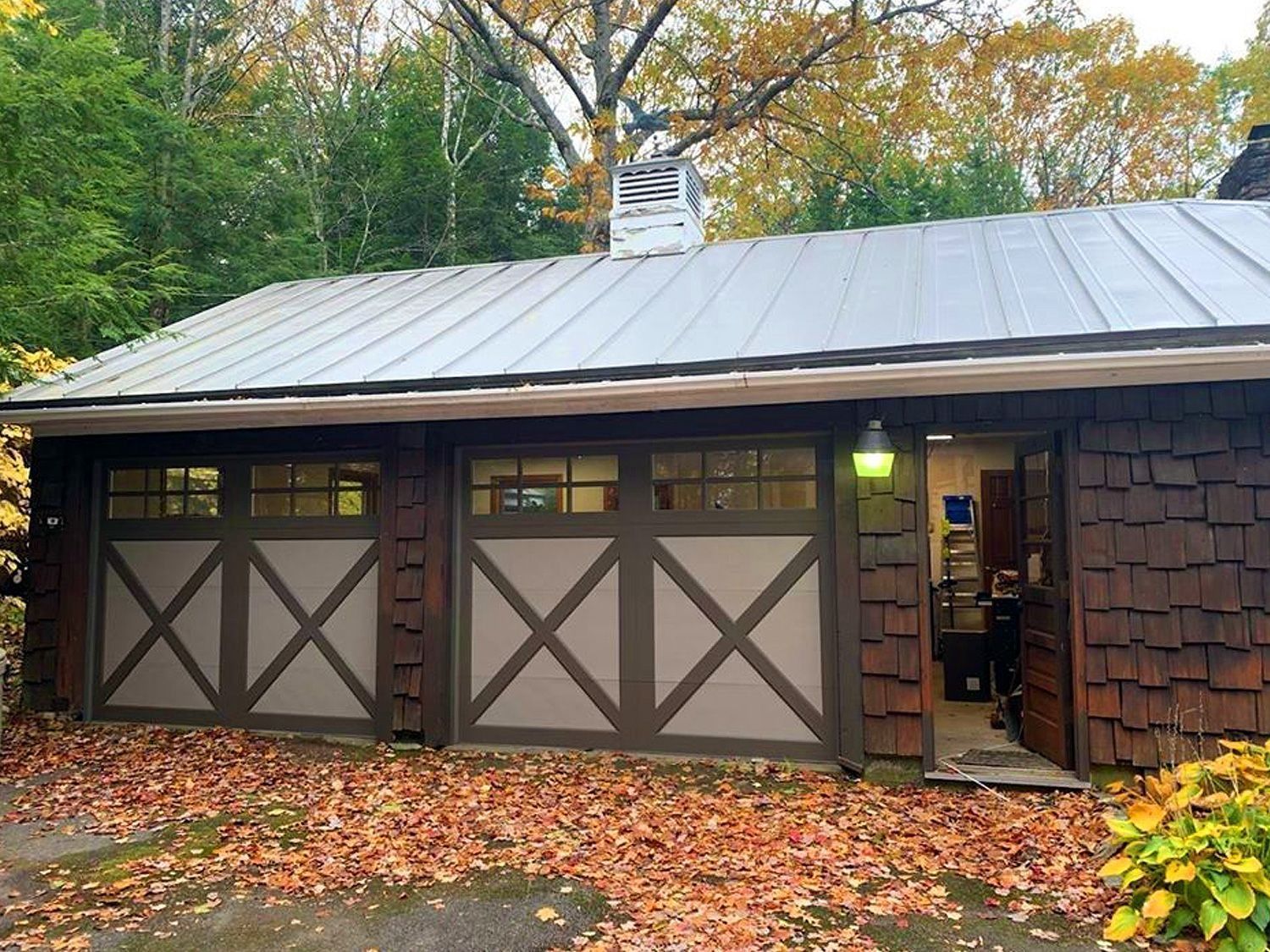 Two-car garage with gray metal roof and brown crossbuck doors, framed by autumn trees and fallen leaves.