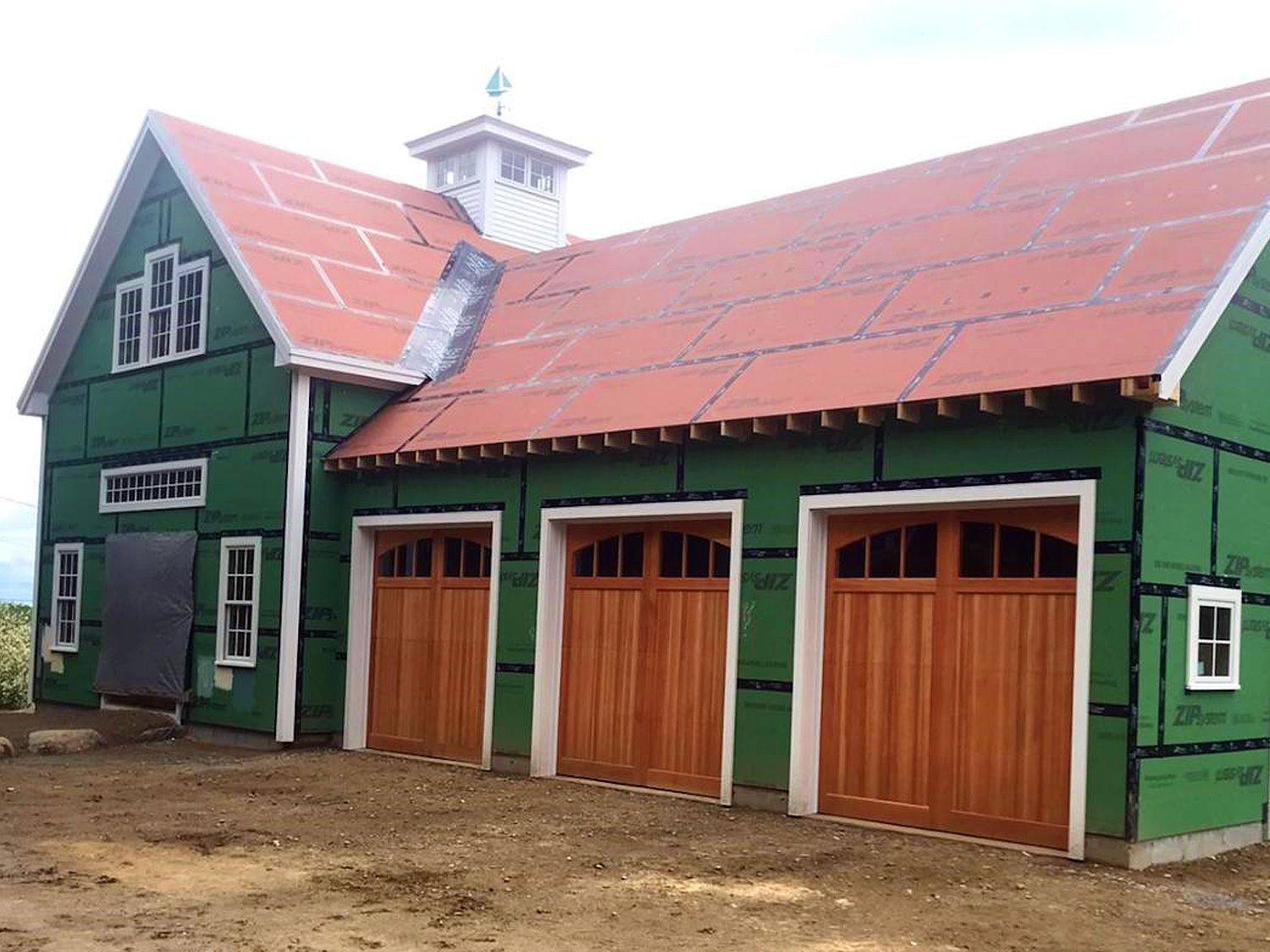 Green barn with red roof and three wooden garage doors in a dirt yard
