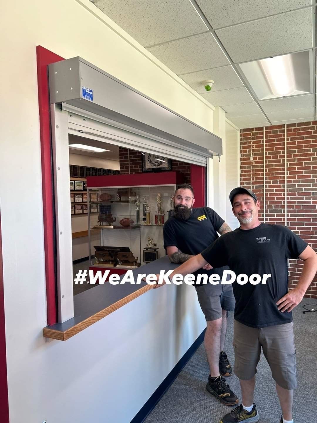 Two men stand beside a service window with #WeAreKeeneDoor overlay on a wall sign.