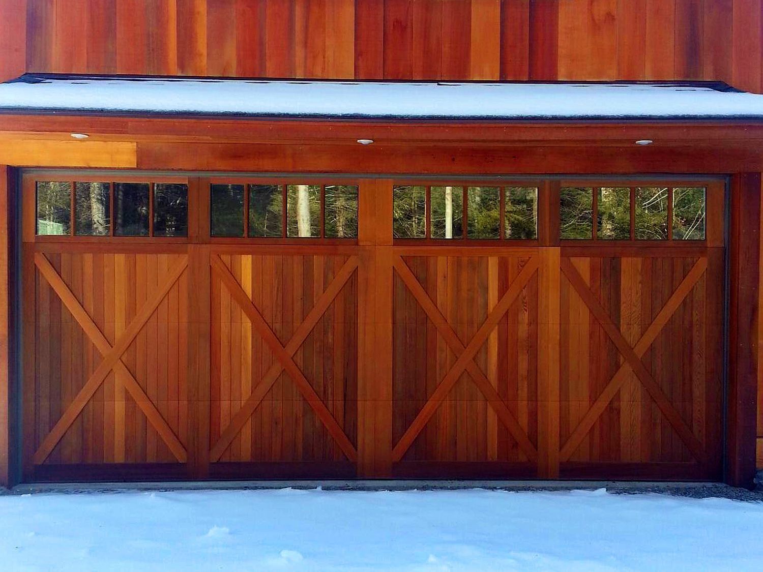 Wooden garage doors with small windows set in a brown building, partly covered by snow.