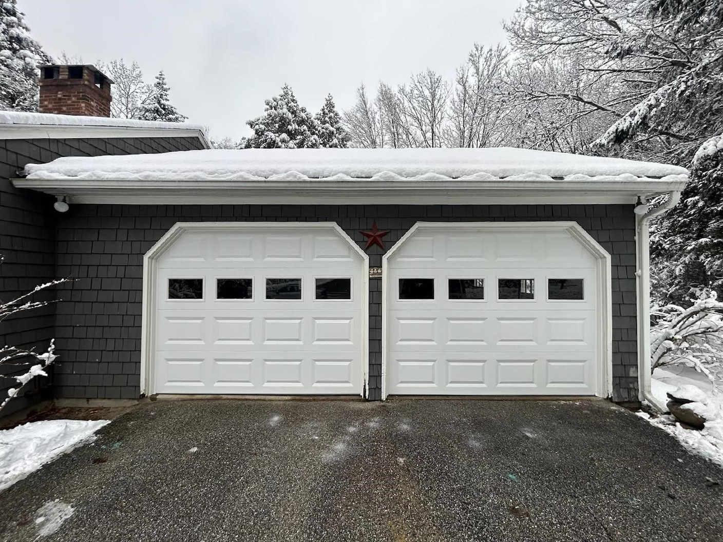 Snow-covered driveway leading to a white double garage with black shutters and a dark brick house