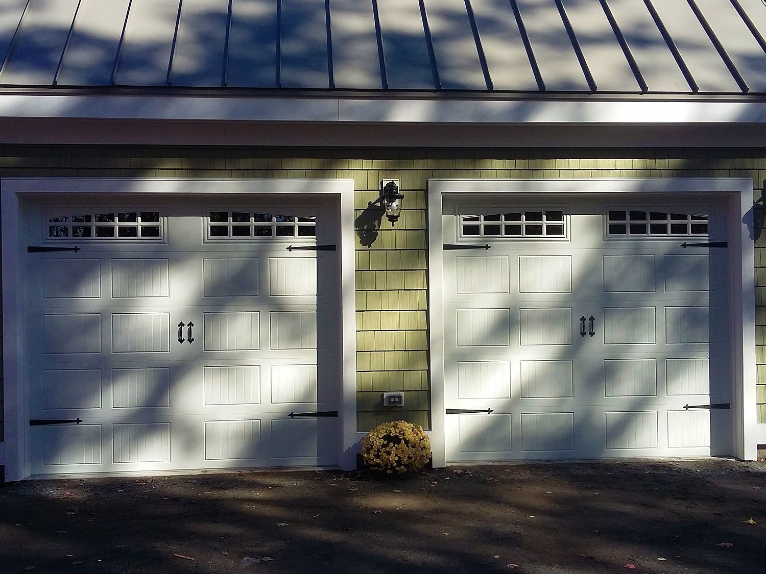 Two white garage doors on a beige house, with a metal roof and sunlight casting shadows across them