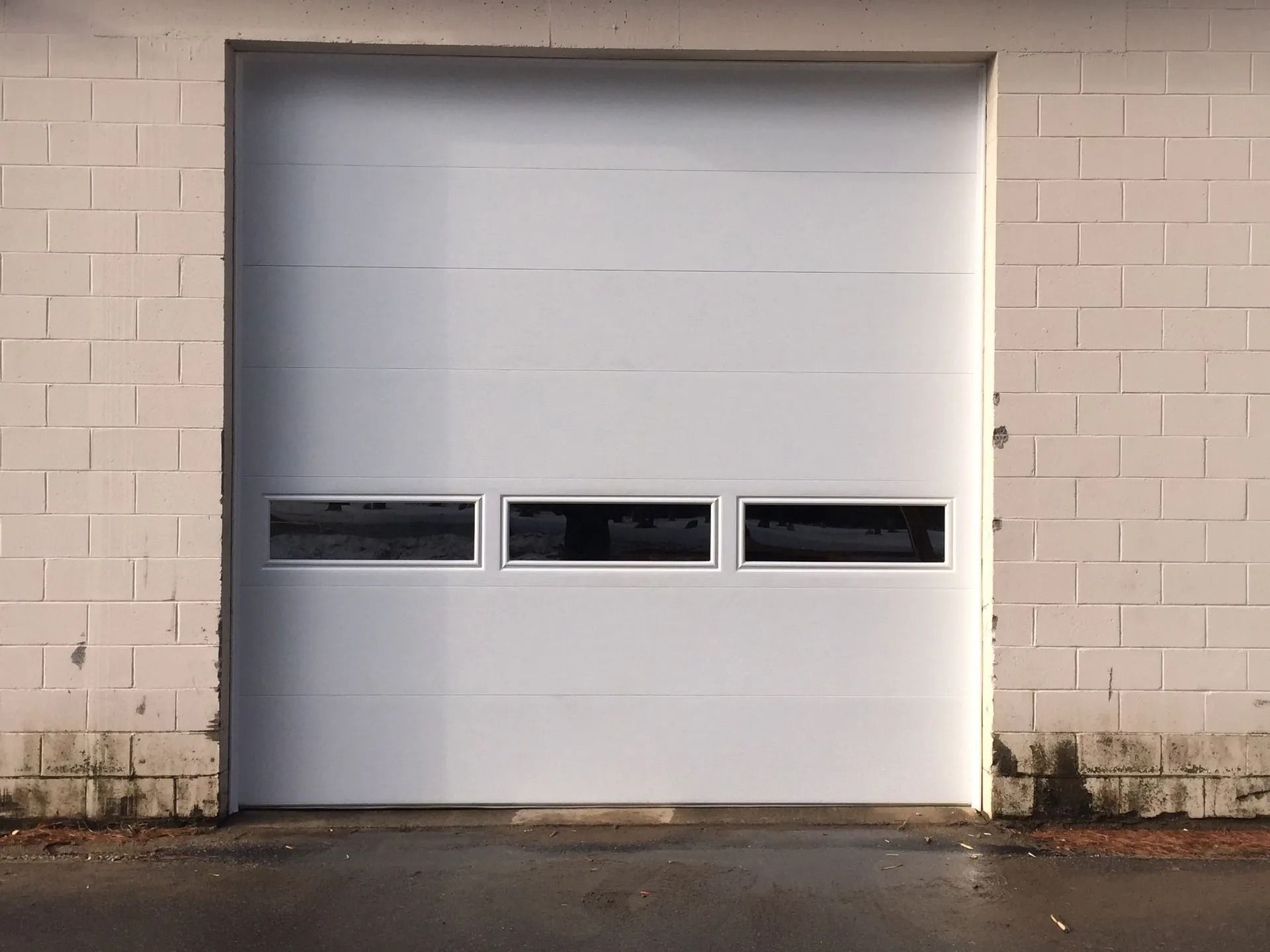 White industrial garage door in a beige brick wall with three small windows near the bottom.