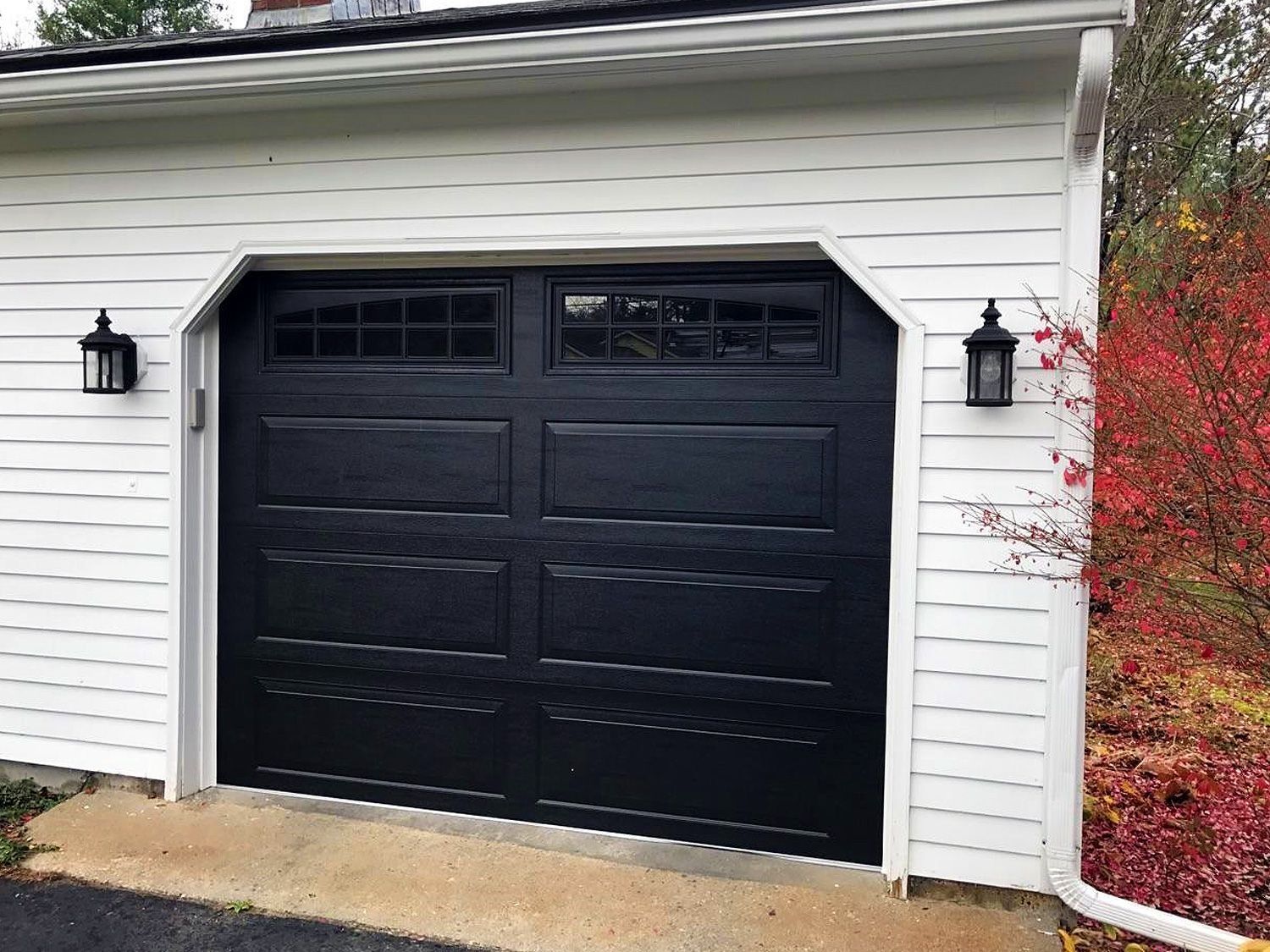 Black double garage door on a white house exterior with wall lanterns and autumn foliage.