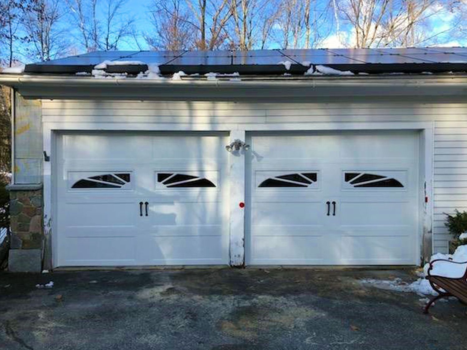 Two white garage doors on a detached garage with snow on the roof and trees in the background