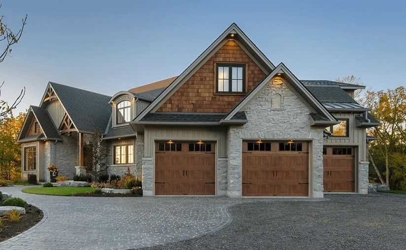 Large stone suburban house with two wooden garage doors, gabled roof, and a gravel driveway at dusk