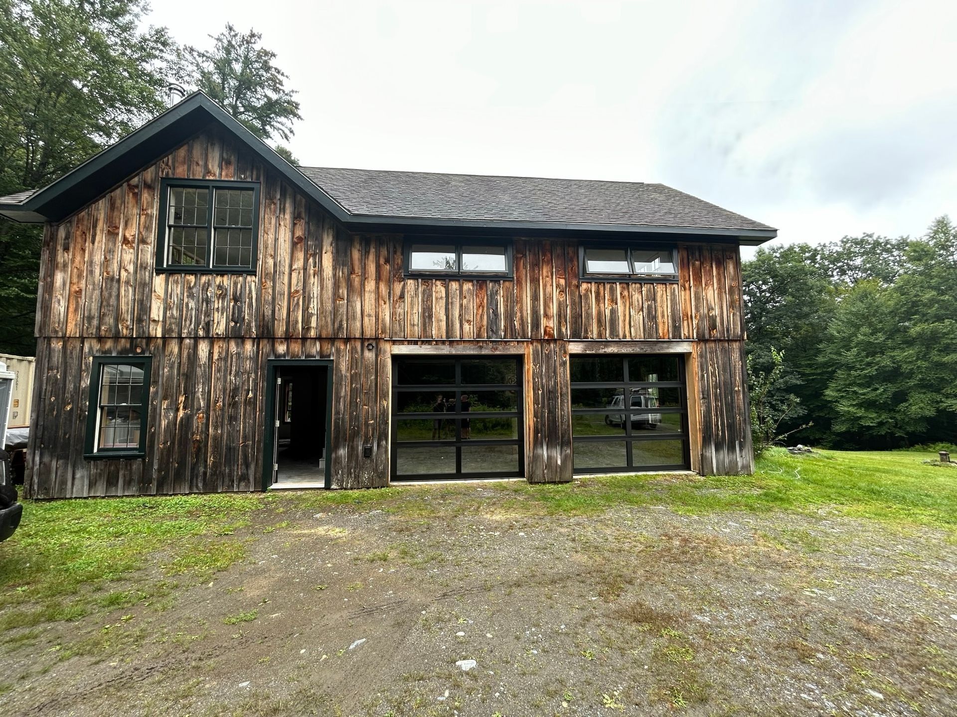 Weathered wooden barn with large open bays and a gravel driveway in front, surrounded by trees