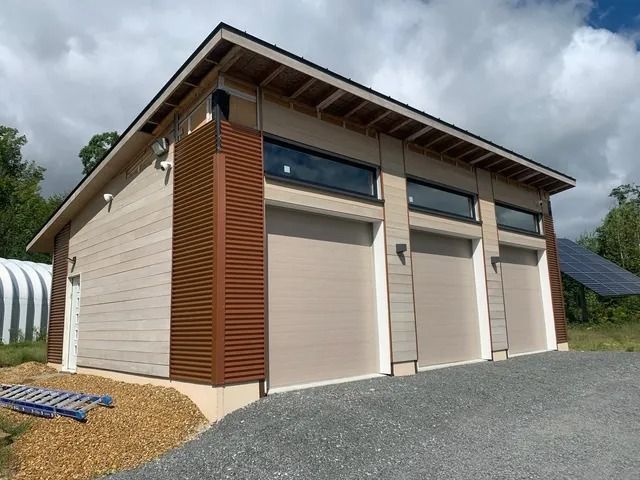 Modern garage with three beige doors and wood trim on a gravel driveway under cloudy skies