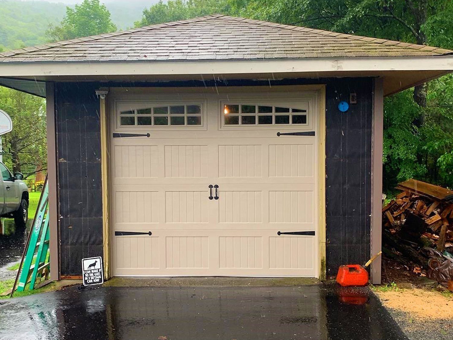 Beige garage door on a dark-sided shed, with wet driveway and green trees in the background.