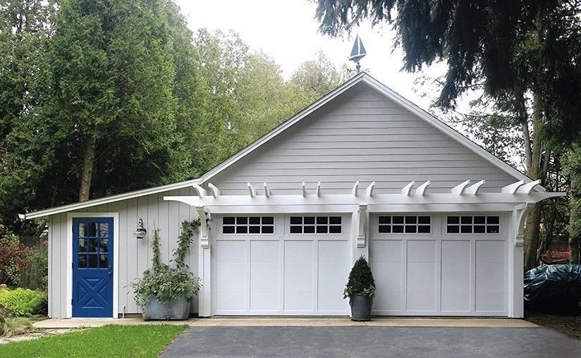 White garage with two doors, blue side door, and gabled roof surrounded by greenery