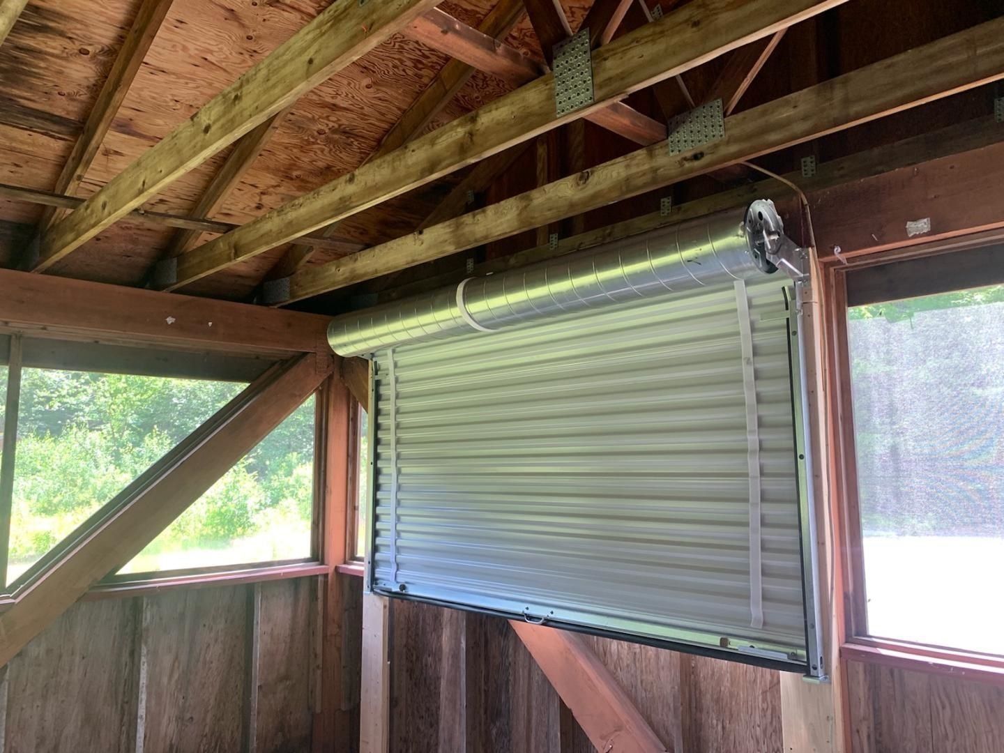 Inside a wooden shed, a metal roll-up window blind is closed over an opening.