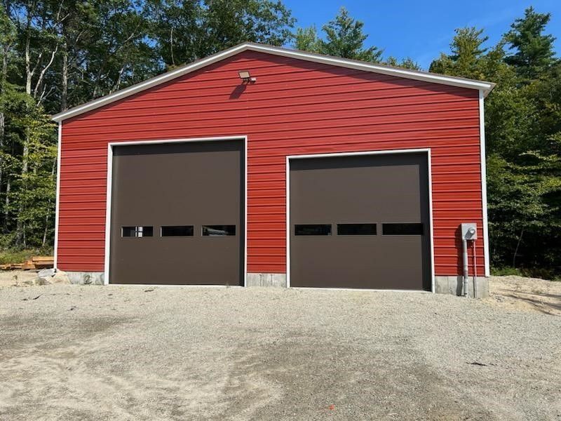 Red garage with two brown doors on a gravel driveway, surrounded by trees.