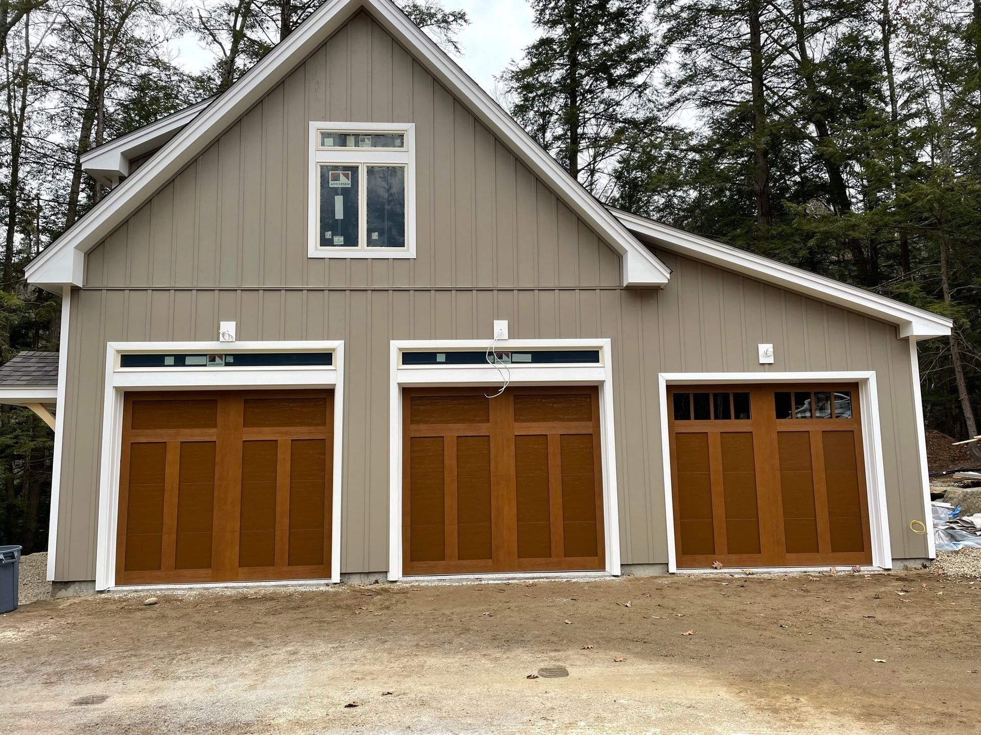 Three-bay garage with brown doors on a beige building, framed by trees and a gravel driveway.