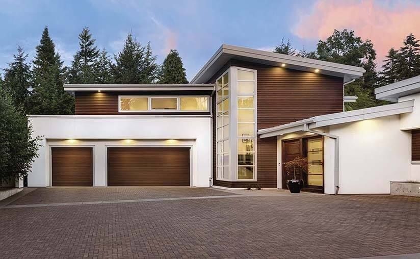 Modern two-story house with wood siding, large windows, and three garage doors at dusk