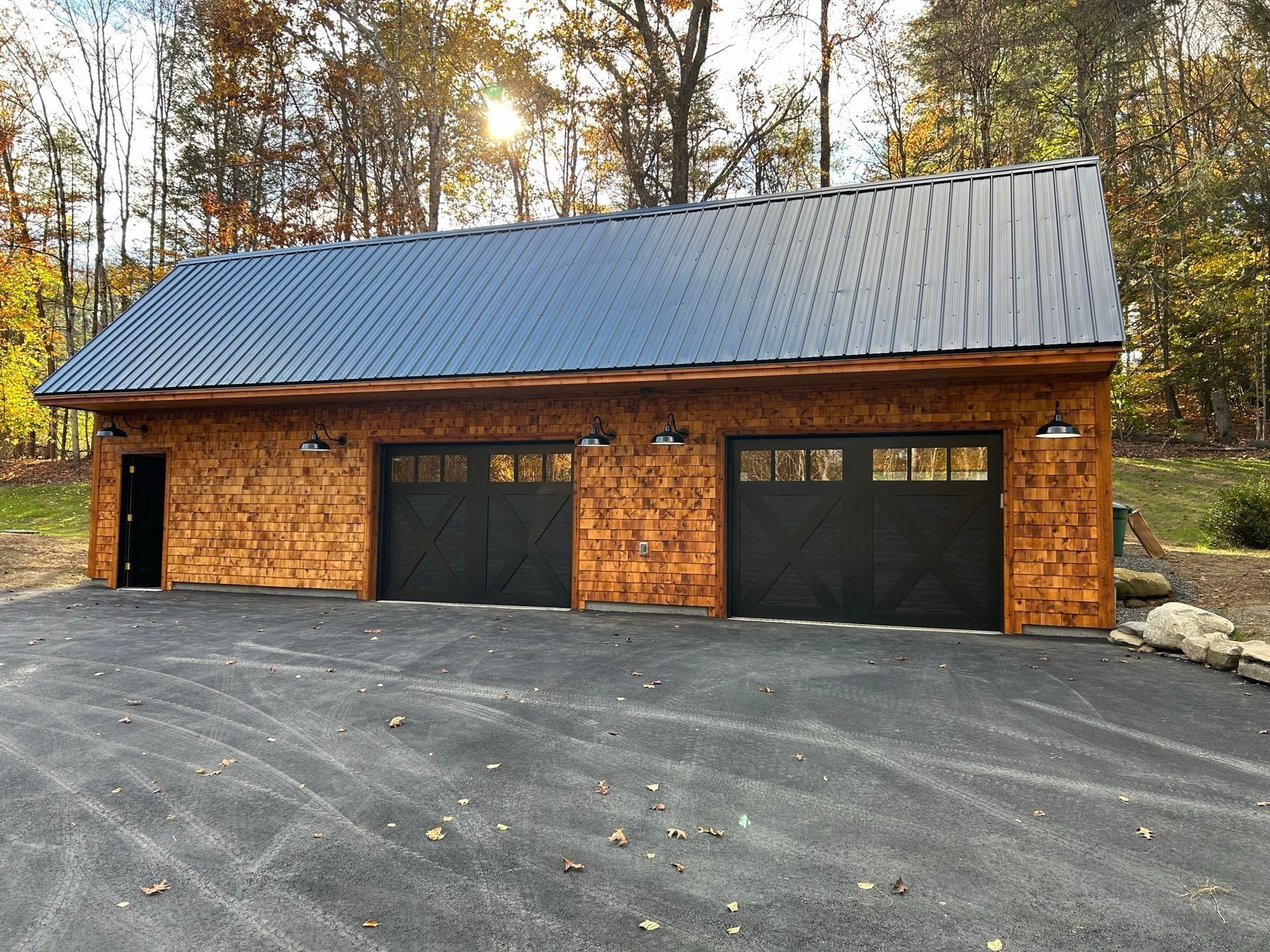 Brick garage with two black doors, a metal roof, and a paved driveway in a wooded setting