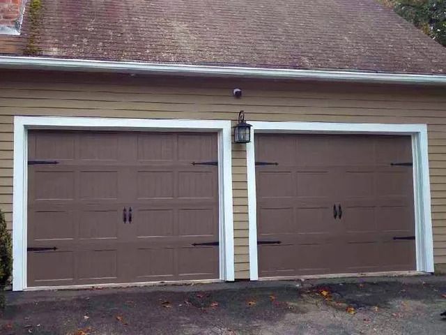 Two brown garage doors with white trim beneath a shingled roof and wall lamp