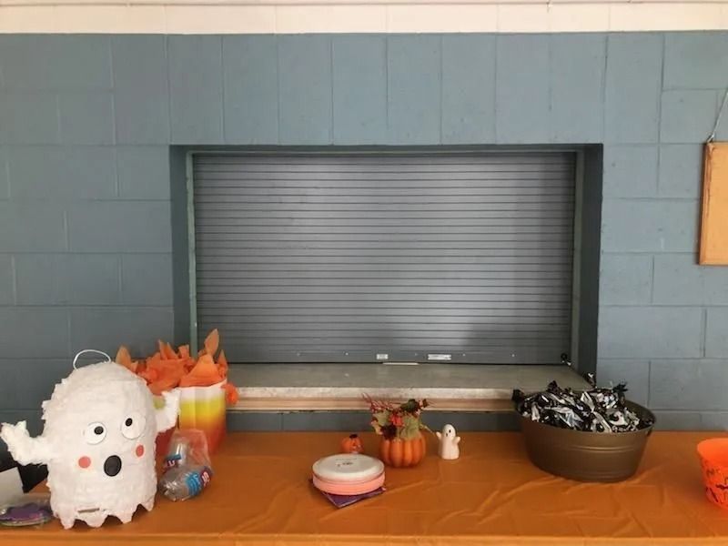 Halloween-decorated table with a white ghost, pumpkins, candy, and a closed service window behind it