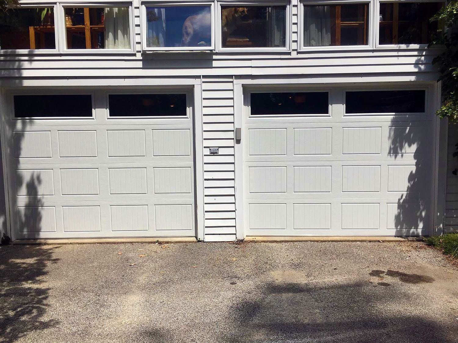 Two white garage doors on a gray house with windows above and a gravel driveway in front