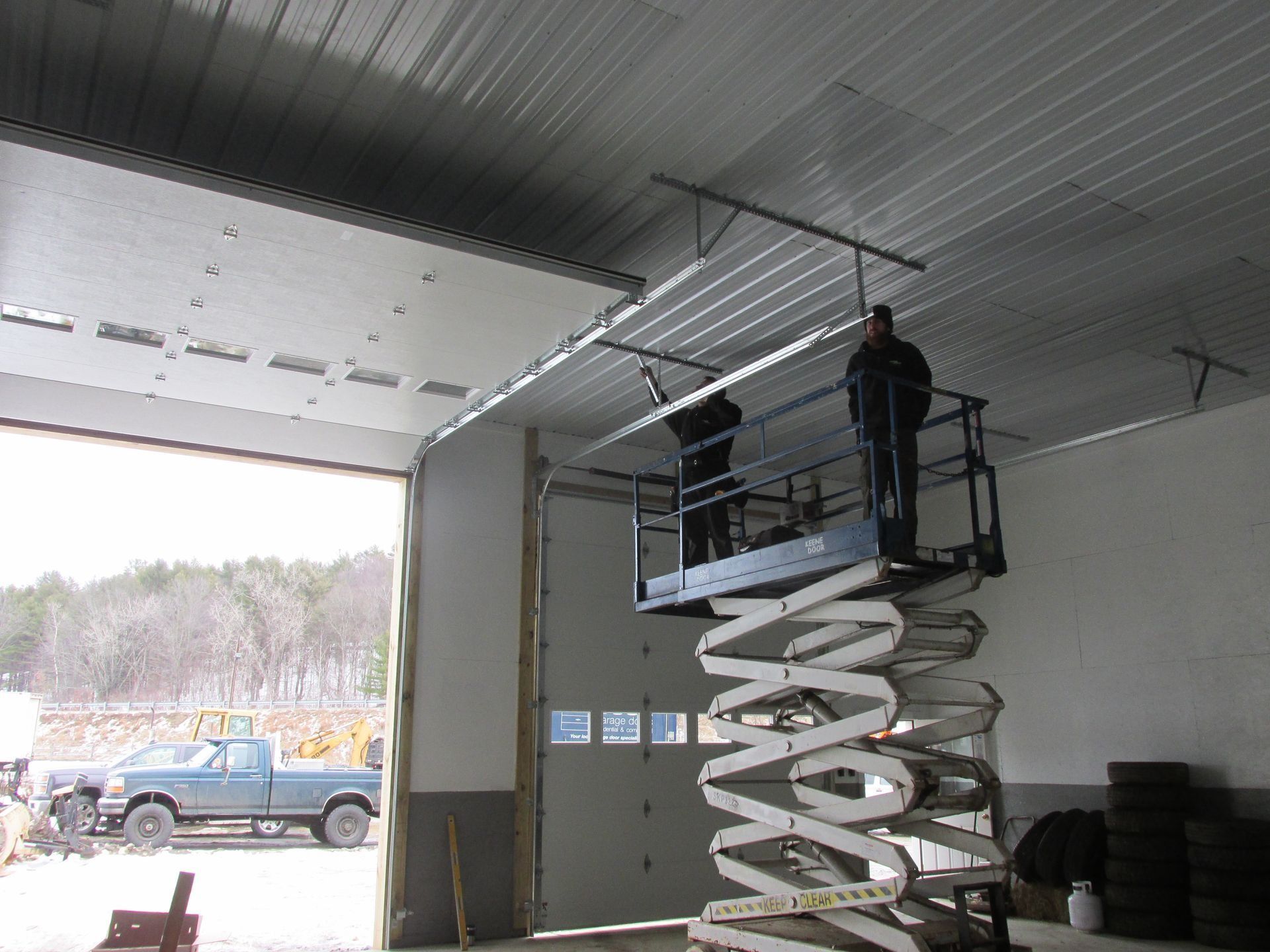 Two workers on a scissor lift inside a garage, raising a large white spiral hose overhead