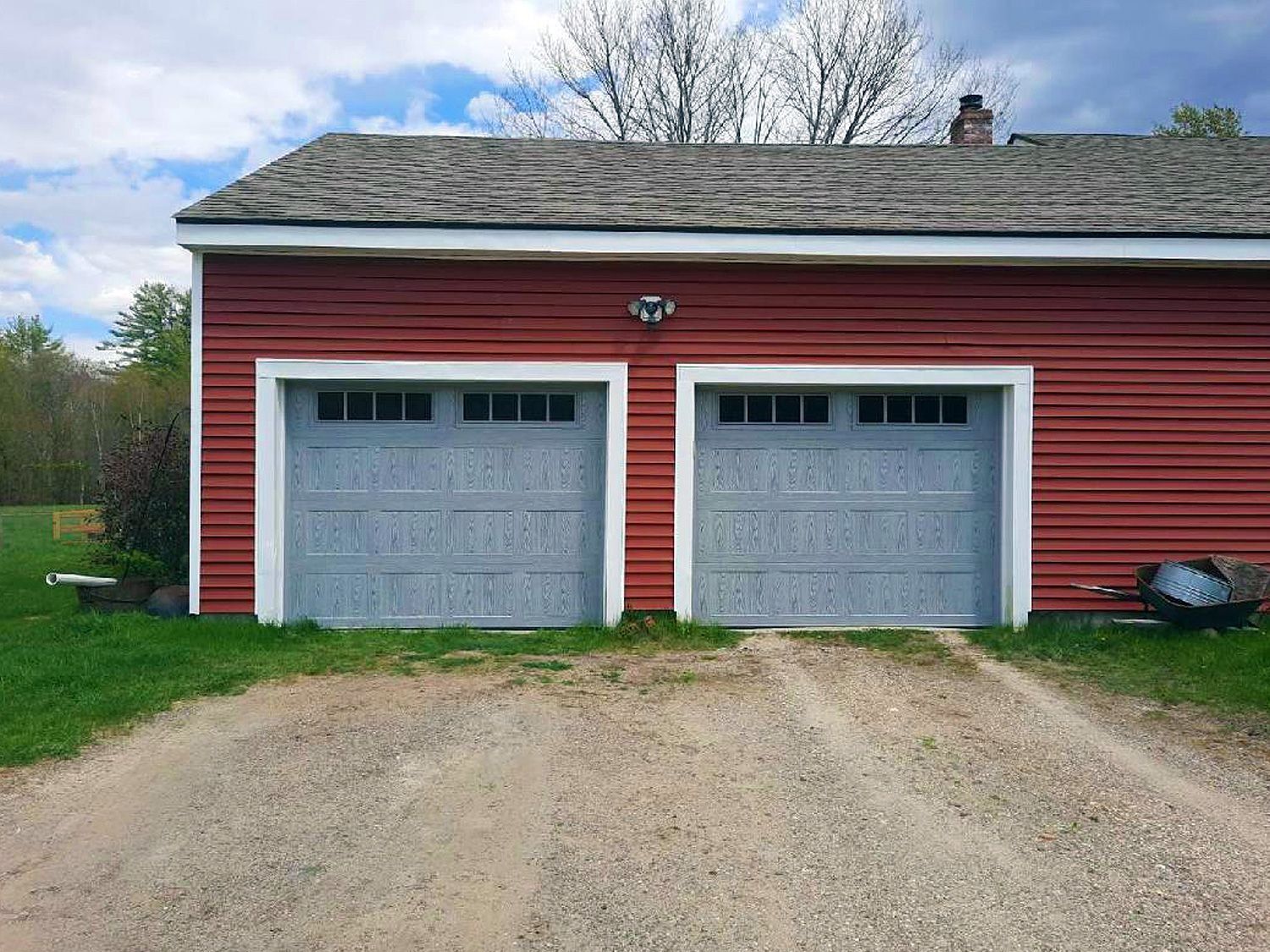 Red garage with two gray doors and a gravel driveway under a partly cloudy sky
