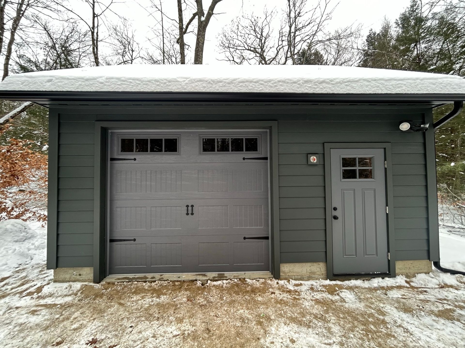 Gray detached garage with a single door, snow on the roof, and a snowy yard.
