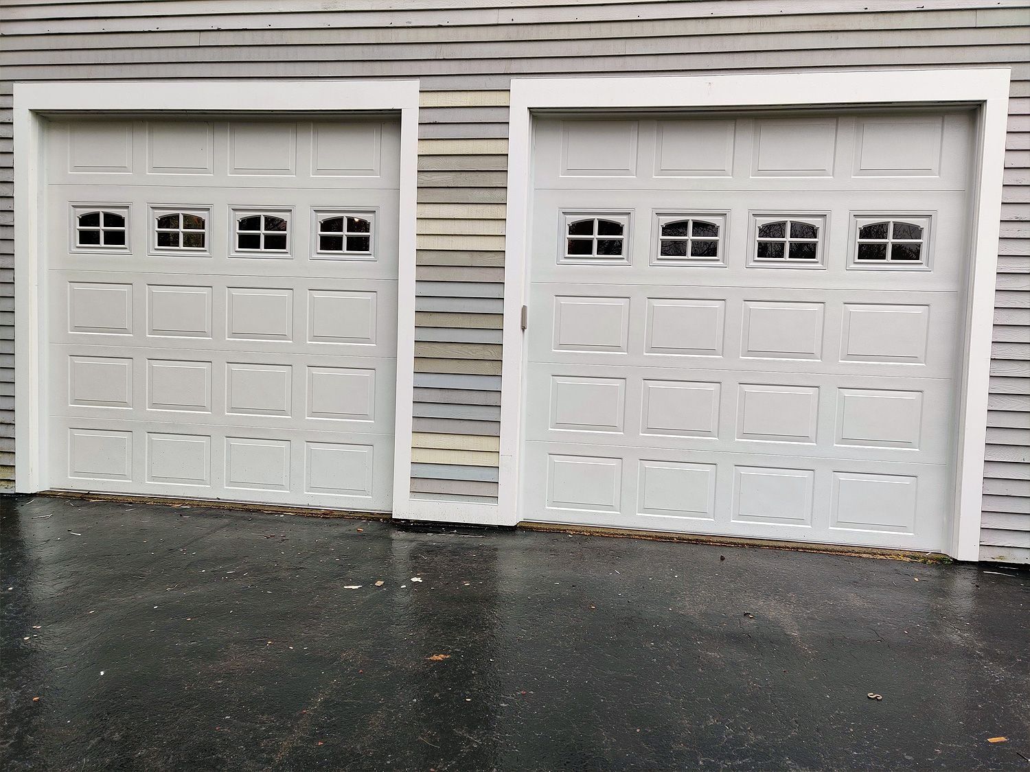 Two white garage doors with small windows on a gray-sided house, seen on a wet driveway.