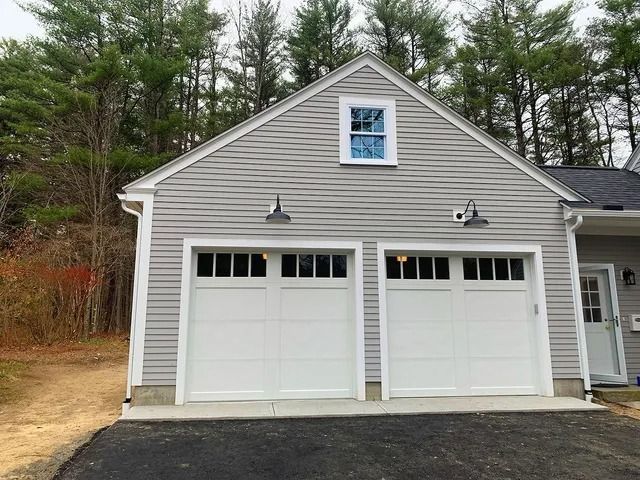 Gray two-car garage with white doors and small upper window beside a wooded driveway