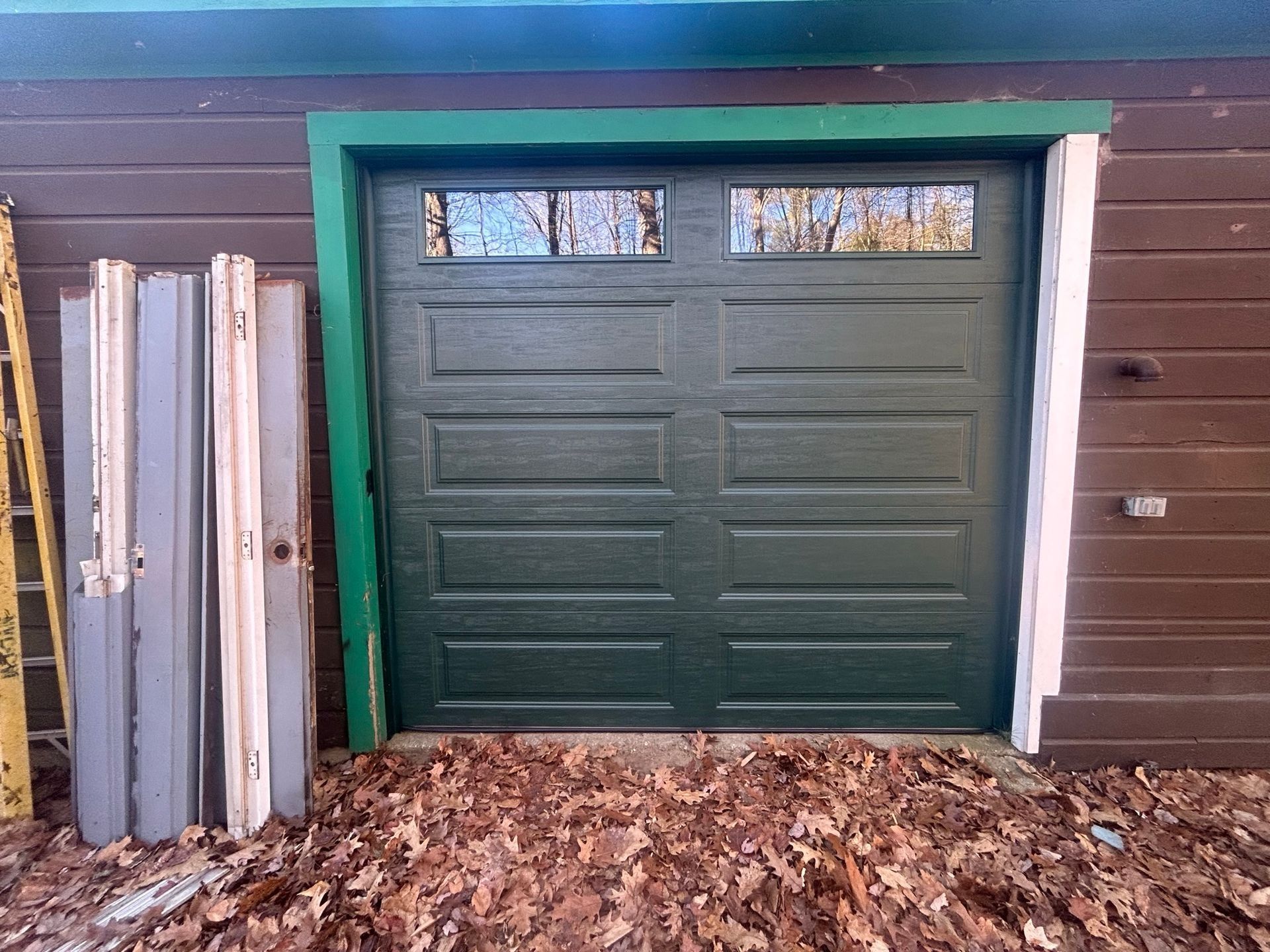 Green garage door in a brown house exterior, with stacked metal panels and leaves on the ground.
