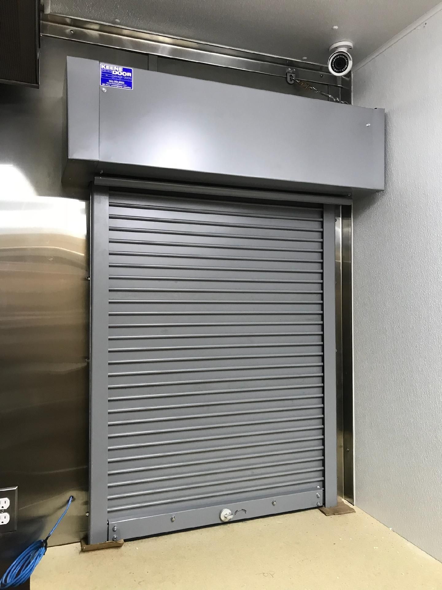Gray metal roll-up door in an indoor industrial hallway, with a large vented unit above it.