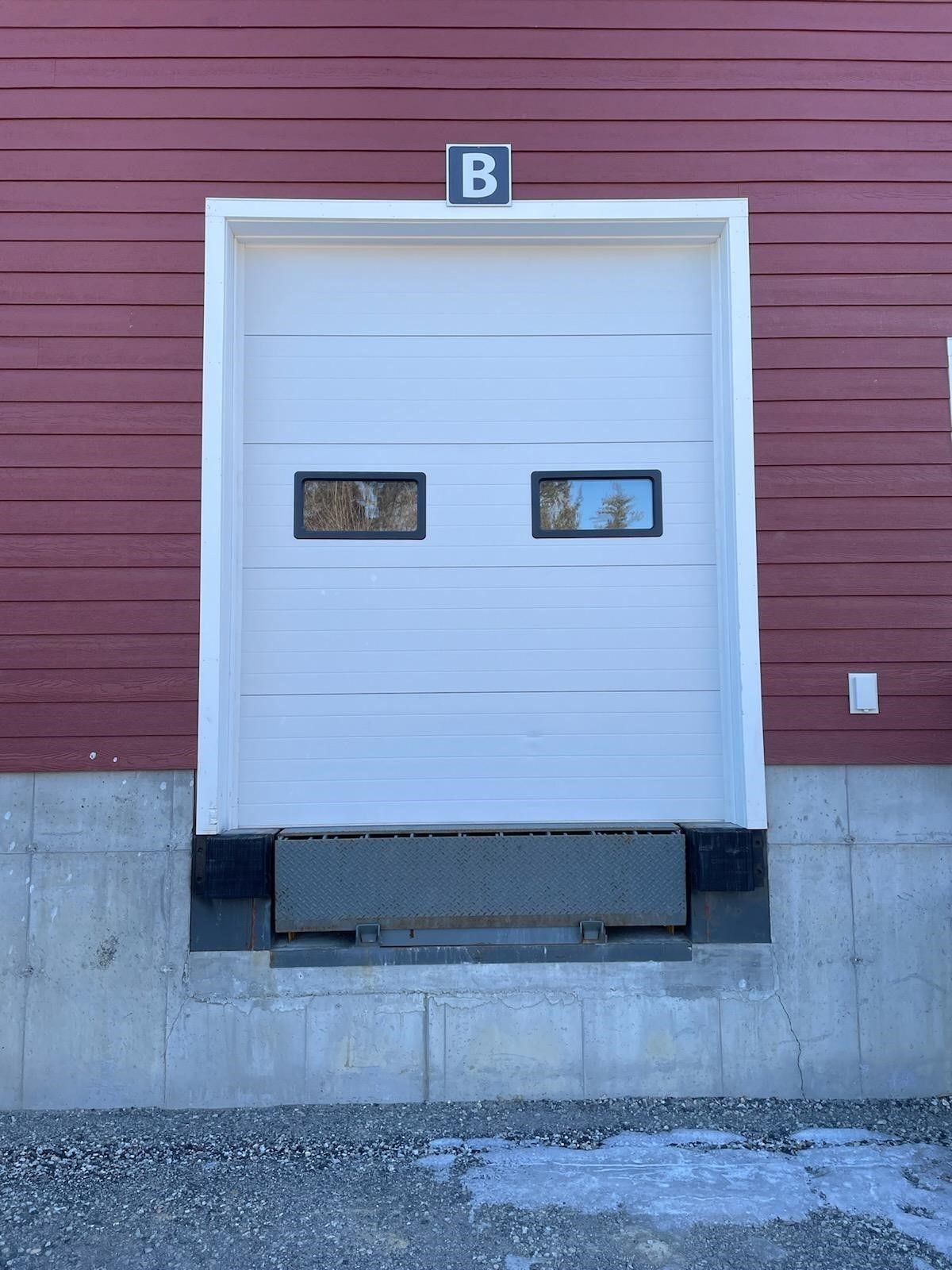 White loading dock door labeled B on a red warehouse wall with concrete base.