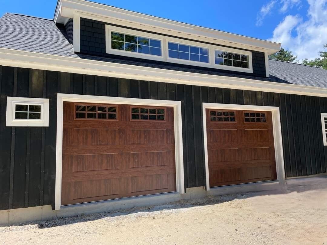 Two wooden garage doors on a black barn-style building with small windows and a white shingled roof