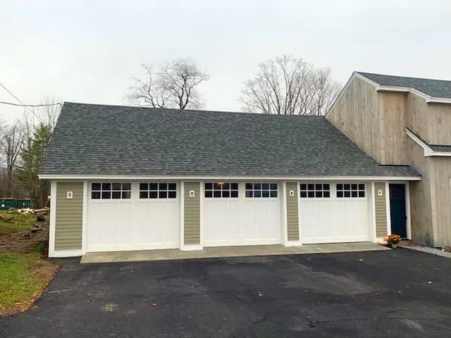Three-car garage with white doors and dark shingled roof beside a house on a paved driveway