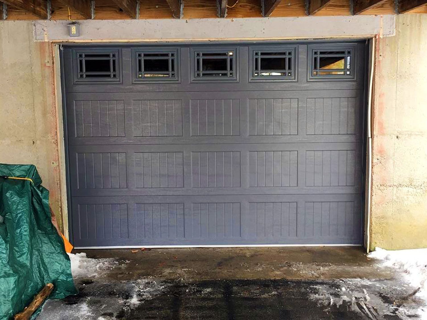 Gray garage door with four small windows, set in a concrete wall with snow on the ground.