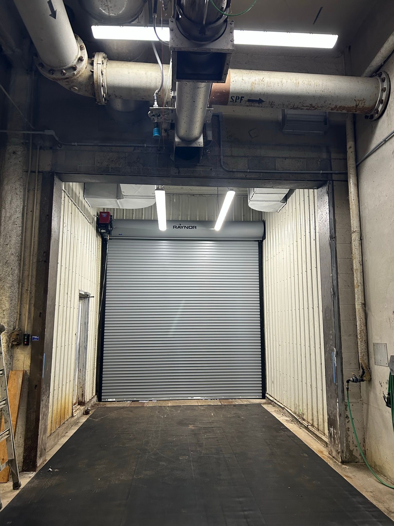 Dim industrial corridor with a closed gray roll-up garage door and overhead pipes, lit by fluorescent lights