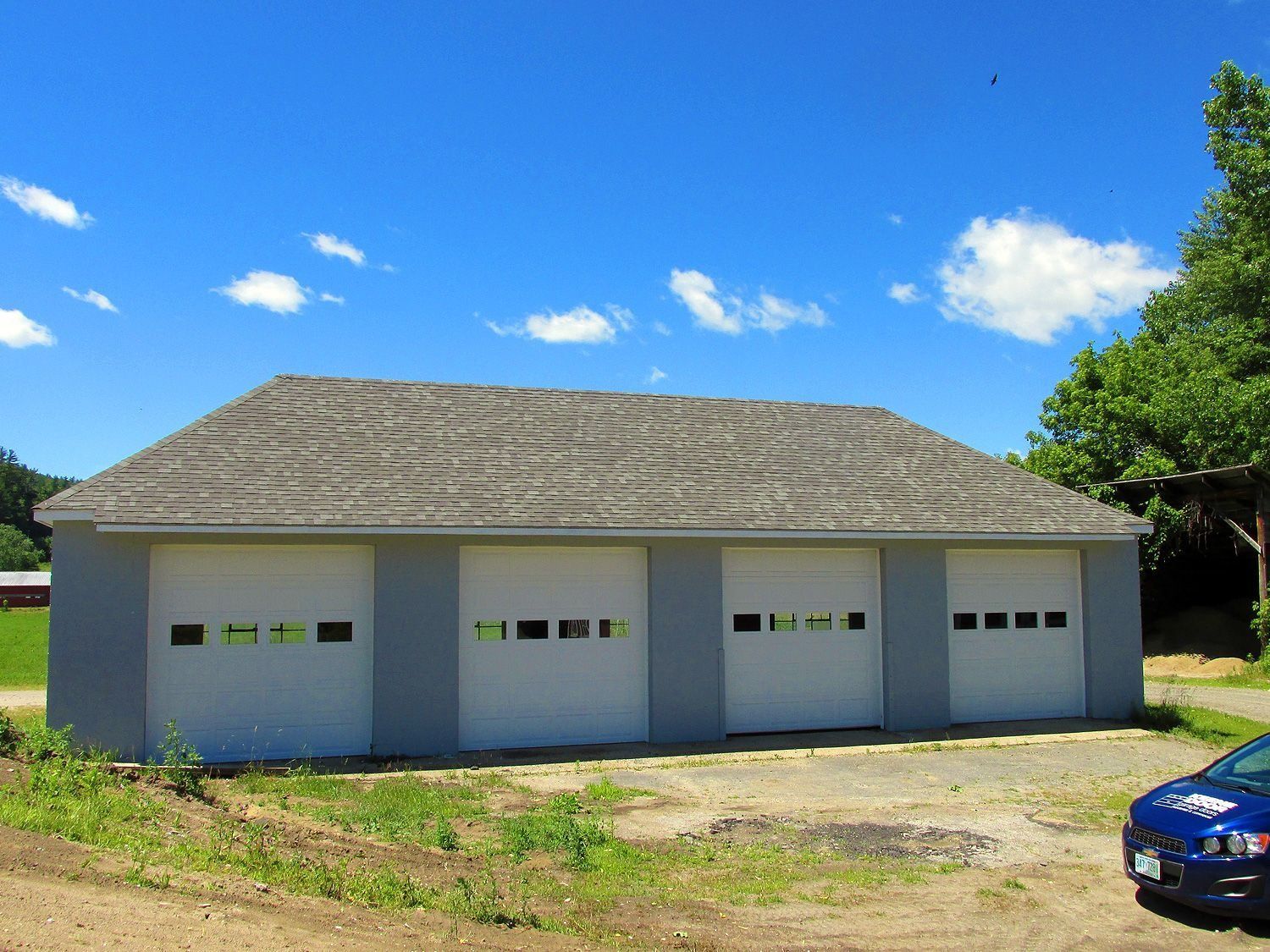 Single-story gray garage with four white doors under a blue sky, with a car at the edge of the frame