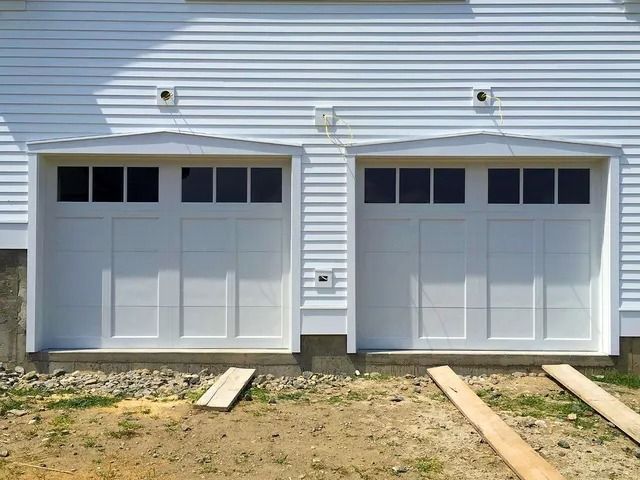 Two white garage doors on a house exterior with siding and a dirt driveway foreground