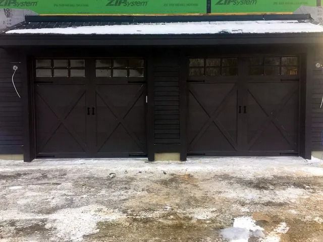 Two dark garage doors on a building under construction, with green sheathing above and snowy ground in front