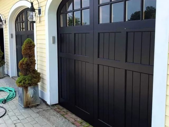 Black paneled garage door on a house exterior beside a potted topiary and garden hose