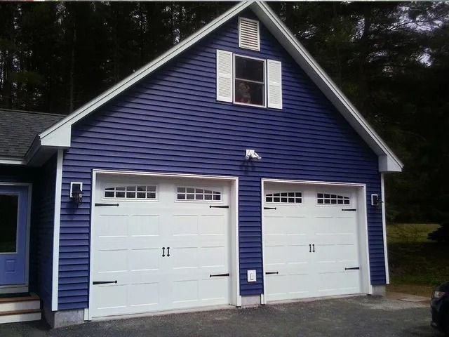 Blue house with two white garage doors and a gabled roof surrounded by trees