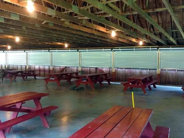 Empty picnic pavilion with red wooden tables under a roof, lit by hanging lights and open sides.
