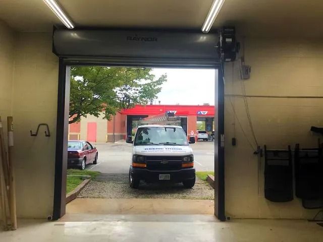 Inside a garage, a white van is parked in an open doorway facing a red-roofed building.