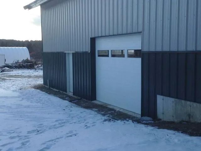 Snowy industrial building with a white garage door and dark siding.